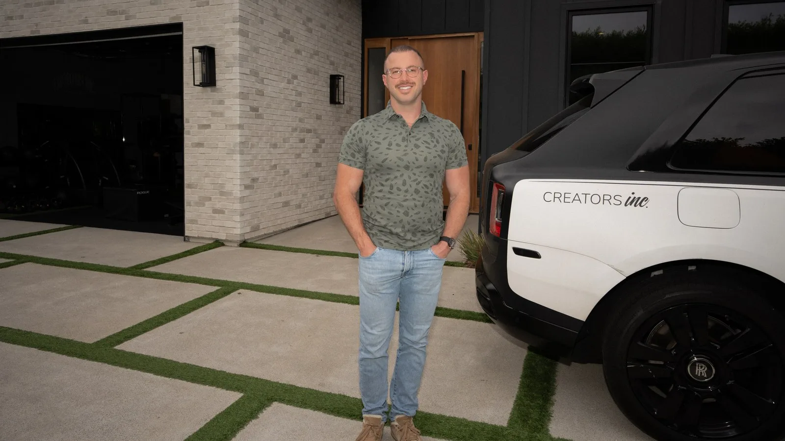 Founder Andy smiling in an olive polo and jeans, standing in a modern driveway next to a Creators Inc branded black and white Rolls-Royce SUV.